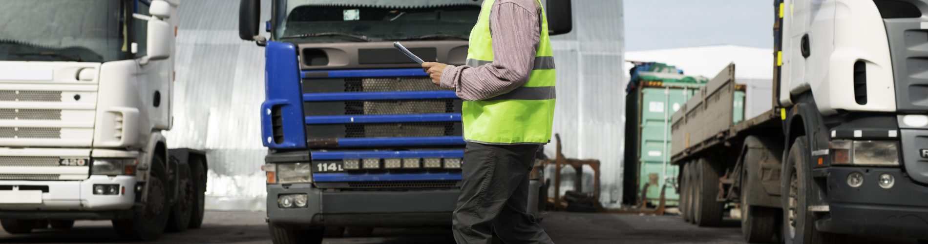 Visão de corpo inteiro de um profissional de logística usando capacete branco e colete verde reflexivo, caminhando por um pátio com vários caminhões alinhados. Ele observa um tablet, representando o inventário operacional e a gestão de ativos em uma frota de grande porte.