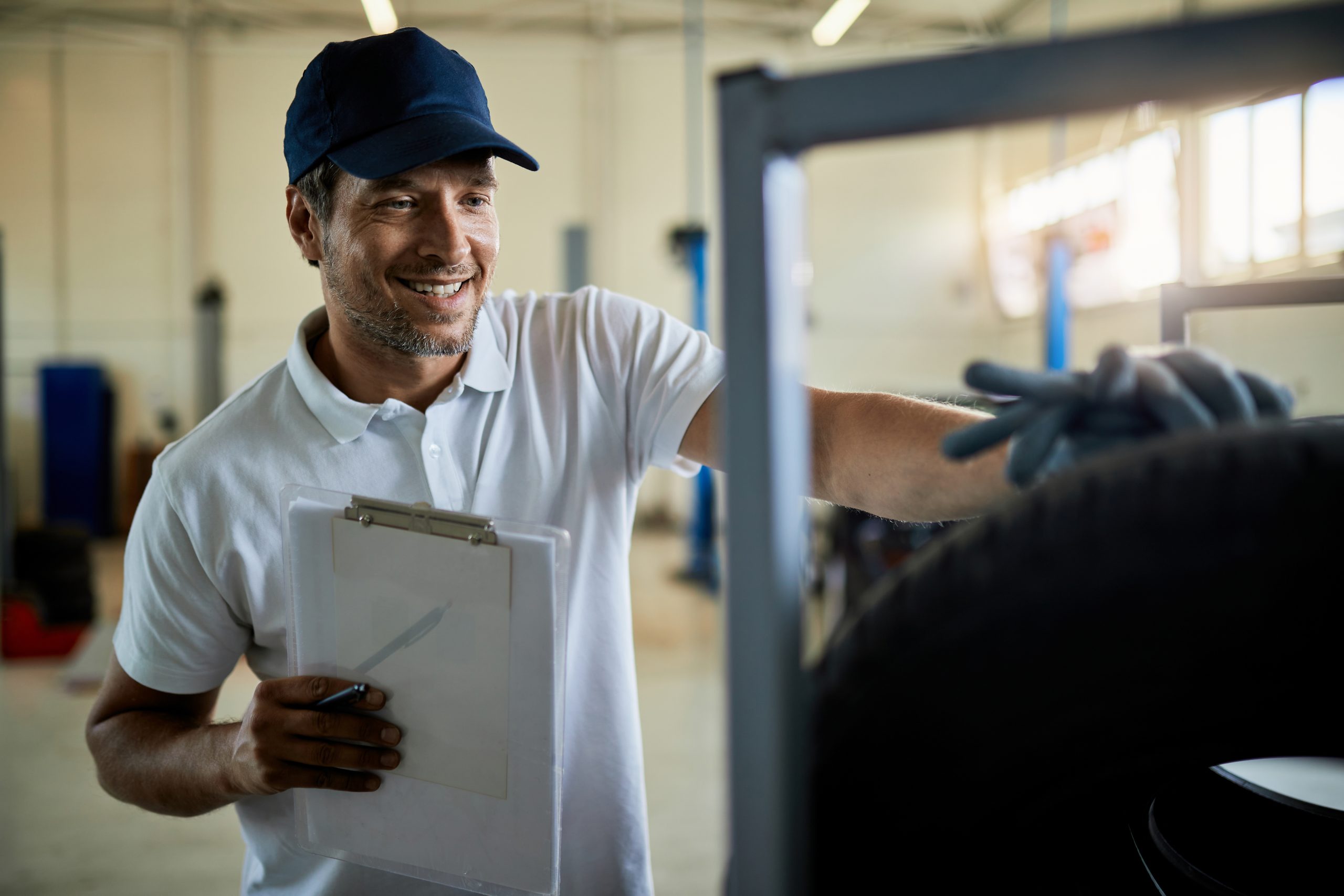 Uma foto horizontal mostra um gestor de frota sorrindo com um boné azul e camisa polo branca, segurando uma prancheta enquanto aponta para o pneu de um carro. Ele tem barba e está olhando para a direita. No fundo, há uma oficina mecânica de borracha e metal. Uma janela à direita brilha com a luz do sol, o pneu do carro está desfocado em primeiro plano, mas a mão direita do mecânico está sobre ele. Sua mão esquerda segura a prancheta branca com a caneta azul.