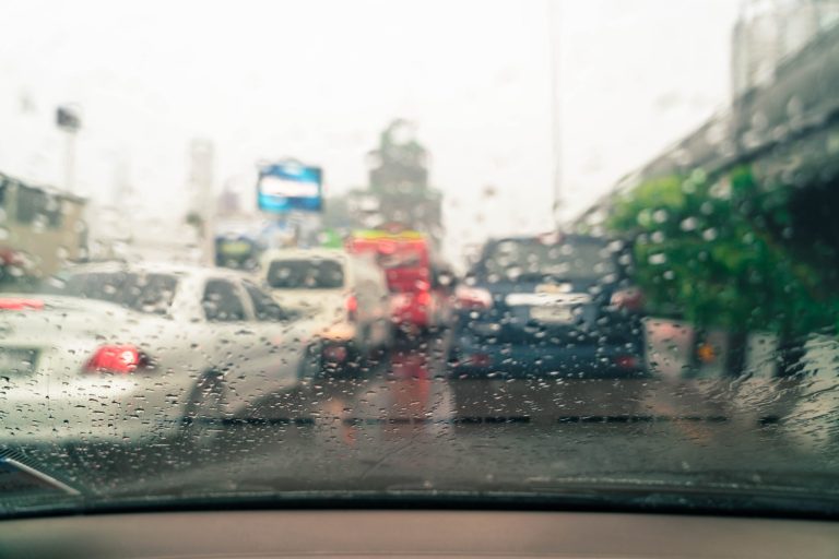 Gotas de chuva no vidro do carro durante condução, reduzindo a visibilidade e destacando riscos na direção e na frenagem em pista molhada.