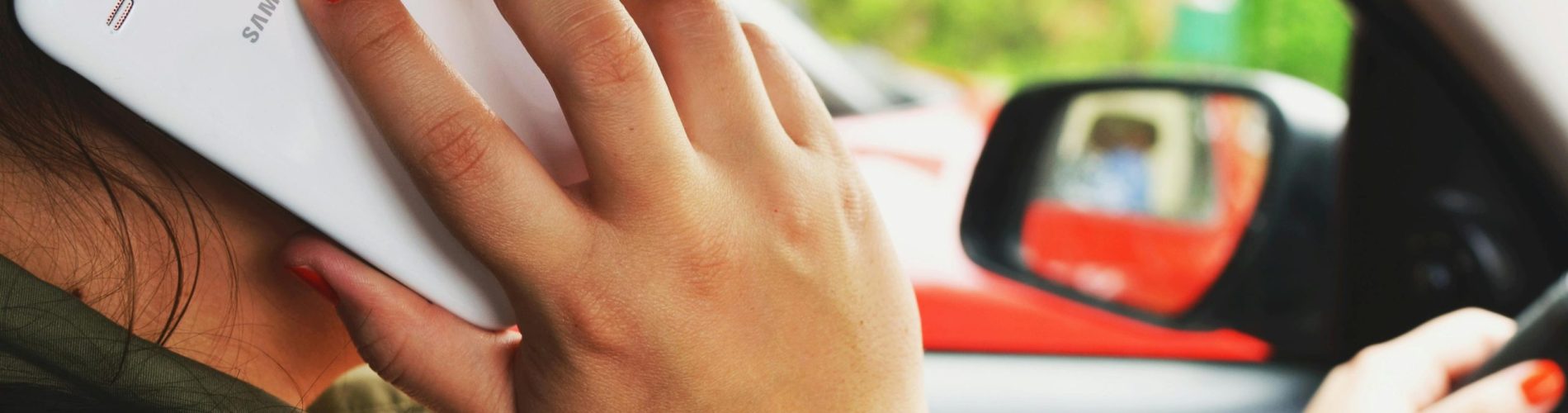 Close-up do interior de um automóvel em movimento, mostrando as mãos de um motorista segurando um smartphone sobre o volante. A imagem foca no perigo da distração visual e manual ao dirigir em uma estrada.