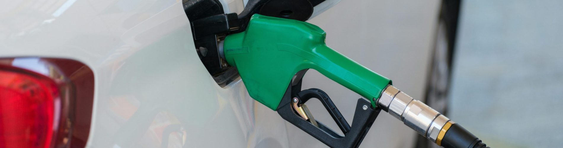 Fotografia em grande plano de um bocal de bomba de combustível preto inserido no tanque de um carro branco durante o reabastecimento num posto de gasolina. A imagem foca no detalhe da mangueira e da entrada de combustível, simbolizando o abastecimento corporativo e a gestão de despesas com combustível.