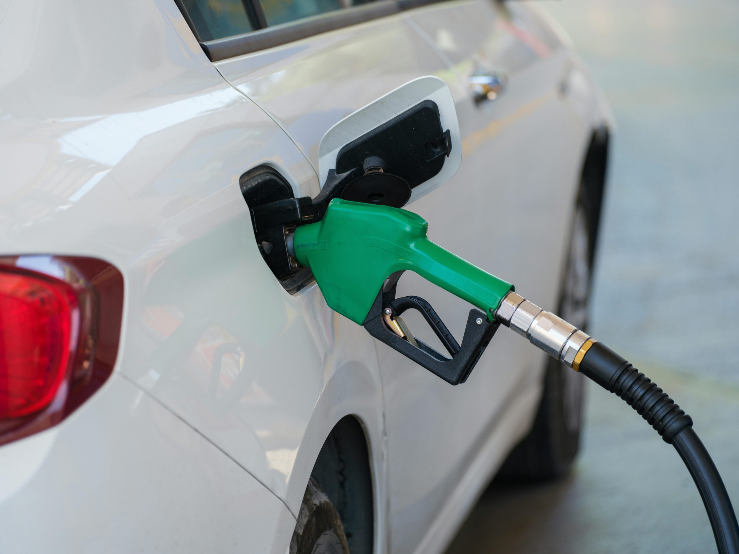 Fotografia em grande plano de um bocal de bomba de combustível preto inserido no tanque de um carro branco durante o reabastecimento num posto de gasolina. A imagem foca no detalhe da mangueira e da entrada de combustível, simbolizando o abastecimento corporativo e a gestão de despesas com combustível.