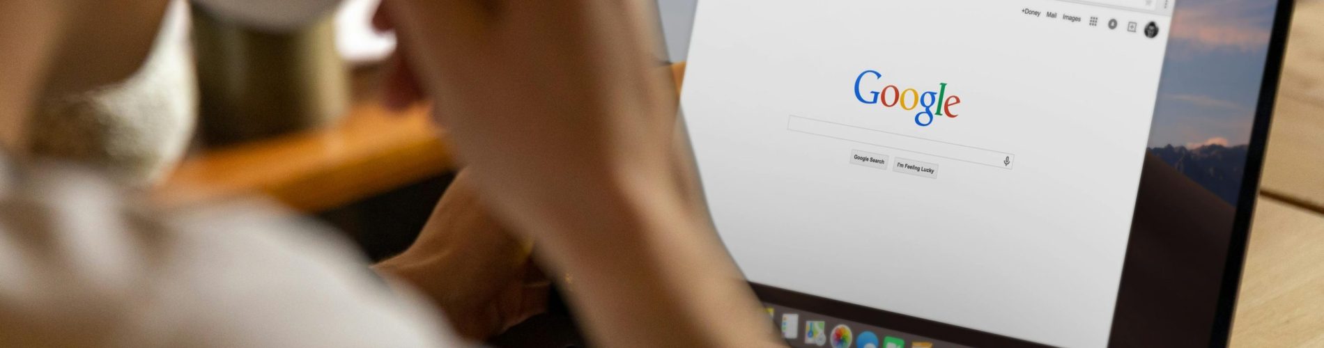 Uma fotografia com foco seletivo em plano detalhe mostrando um ambiente de trabalho organizado e minimalista. No lado esquerdo, em primeiro plano, há uma xícara de café de cerâmica branca com café preto, posicionada sobre um pires branco. Ao lado da xícara, à direita, encontra-se um notebook prateado aberto, exibindo uma tela com gráficos e dados borrados ao fundo. Entre o café e o computador, há um bloco de notas (notebook) de capa escura com uma caneta preta pousada sobre ele. A iluminação é suave e natural, sugerindo um ambiente de escritório ou home office produtivo.